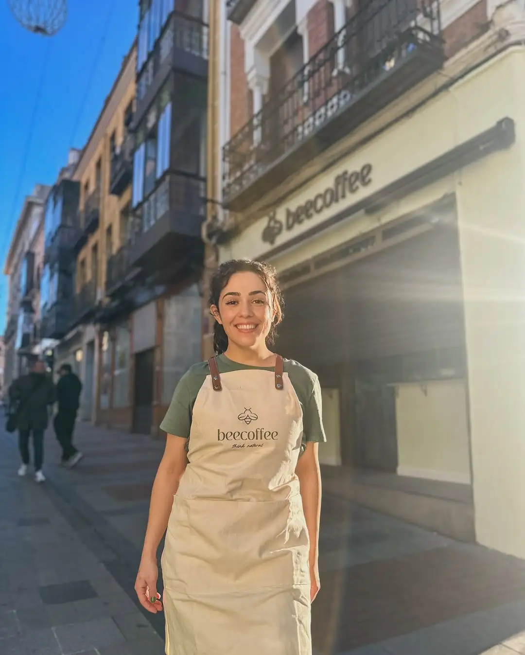 Fotografía en primer plano de María, barista y fundadora de Beecoffee, con rostro sonriente. Está situada en la calle y tiene justo detrás la entrada del local.